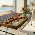 A professional glass desk with a dark walnut wood panel on the left, overlooking a sunny Sunshine Coast beach. On the desk, an oak abacus and a document titled "Our Transparent Fee Structure" sit in the foreground, bathed in warm morning light.
