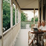 A soulful, film-shot photograph of a 60-year-old daughter and her 90-year-old father sitting on a traditional Queenslander veranda, looking at a tablet together with a sense of security and family protection.