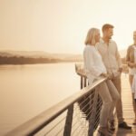 A diverse multi-generational family—an older couple, a middle-aged daughter, and two young children—standing together on a sunlit boardwalk by a river at sunset, representing secure financial legacy and retirement.