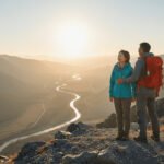 An active East Asian couple in their 60s standing on a mountain peak at sunset, looking out over a winding river valley, representing financial freedom and peace of mind.