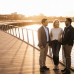 A diverse group of three professionals and retirees in their 60s talking on a modern sun-drenched boardwalk by a river, symbolising community, trust, and a clear path to financial freedom.