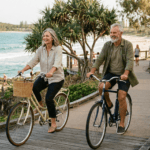 A happy man and woman in their 60s riding vintage-style bicycles along the Mooloolba boardwalk, with pandanus trees and the Pacific Ocean in the background.