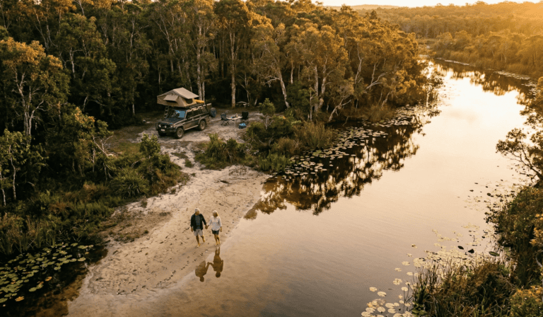 How Much Money Do I Actually Need to Retire on the Sunshine Coast? (2026 Guide) 4 High-angle drone shot of an older couple walking barefoot on a sandy riverbank next to a 4WD camper in the Noosa Everglades at sunset. How Much Money Do I Actually Need to Retire on the Sunshine Coast? (2026 Guide).