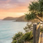 A happy retired couple in their 50s walking hand-in-hand along the Noosa National Park boardwalk at sunrise on the Sunshine Coast. They are looking out over the calm ocean horizon, framed by coastal Pandanus trees in warm, golden morning light. Retirement Planning Sunshine Coast.